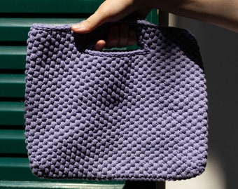 May include: A small, lavender-colored crocheted tote bag with a textured, puffy design. The bag has a single handle and is held in a hand.