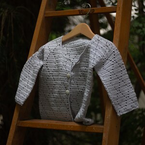 May include: A light gray, crocheted baby cardigan with long sleeves, hanging on a wooden hanger. The cardigan has a button-up front with two buttons. The garment is displayed on a wooden ladder, with a blurred green background.