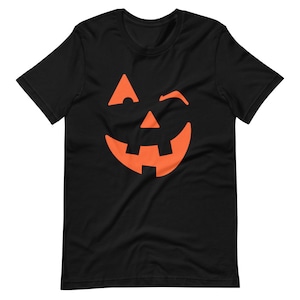 Puede incluir: Camiseta negra con un gr&aacute;fico de cara de calabaza naranja gui&ntilde;ando un ojo.