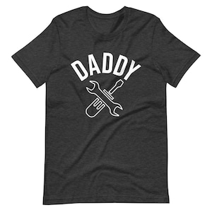 K&ouml;nnte beinhalten: Dunkelgraues T-Shirt mit einem wei&szlig;en Grafikdruck eines Schraubenschl&uuml;ssels und Schraubendrehers, die sich kreuzen, mit dem Wort "DADDY" dar&uuml;ber.