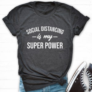 Puede incluir: Camiseta gris oscuro con el texto blanco "SOCIAL DISTANCING is my SUPER POWER". La camiseta es de cuello redondo y parece estar hecha de un material suave. Se ve un par de zapatillas altas negras con suelas y cordones blancos.