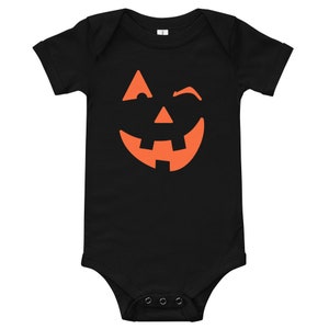 Puede incluir: Mono de beb&eacute; negro con un gr&aacute;fico de cara de calabaza naranja. La cara de calabaza est&aacute; sonriendo y gui&ntilde;ando un ojo.