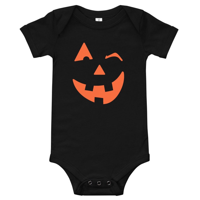 Puede incluir: Mono de beb&eacute; negro con un gr&aacute;fico de cara de calabaza naranja. La cara de calabaza est&aacute; sonriendo y gui&ntilde;ando un ojo.