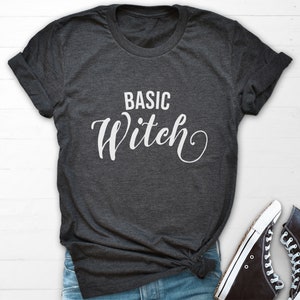 Könnte beinhalten: Ein dunkelgraues T-Shirt mit dem Text "BASIC WITCH" in weißer Kursivschrift.
