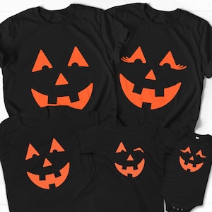 Puede incluir: Conjunto de cinco camisetas negras con caras de calabaza naranja. Las camisetas presentan diferentes expresiones, incluyendo una calabaza gui&ntilde;ando un ojo, una calabaza con pesta&ntilde;as y una cara de calabaza normal.