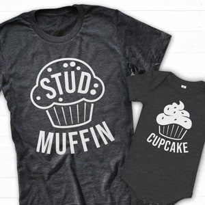 May include: A gray t-shirt with a white cupcake graphic and the text "Stud Muffin". A smaller gray t-shirt with a white cupcake graphic and the text "Cupcake".