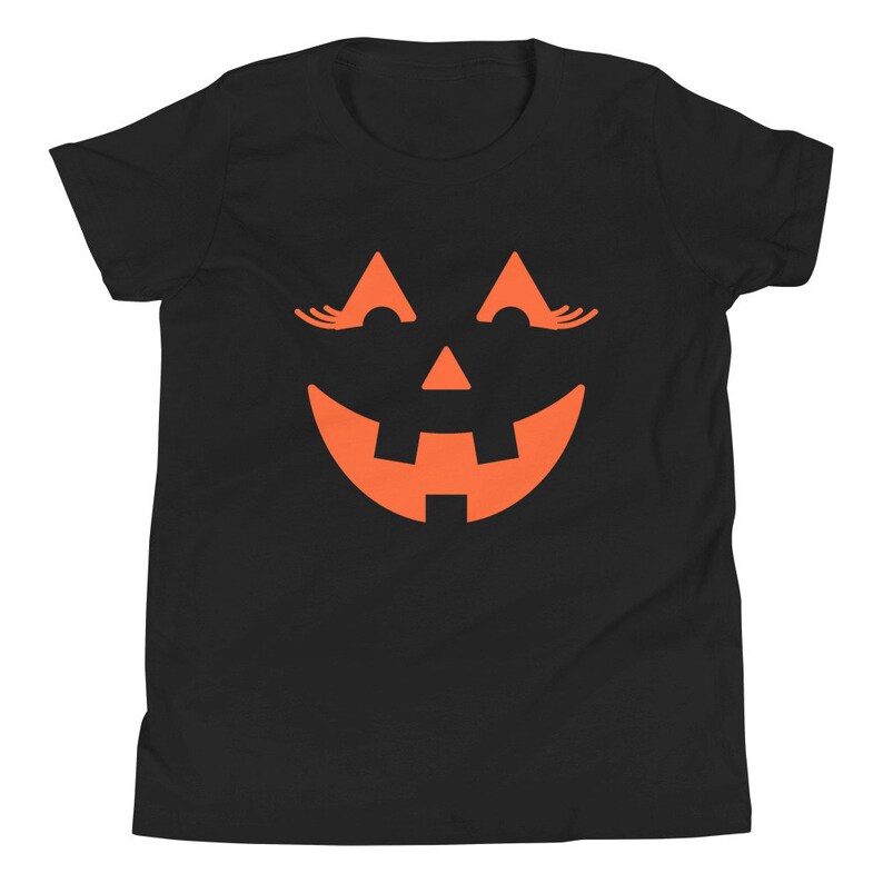 Puede incluir: Camiseta negra para j&oacute;venes con un gr&aacute;fico de cara de calabaza naranja. La calabaza tiene pesta&ntilde;as y una amplia sonrisa.