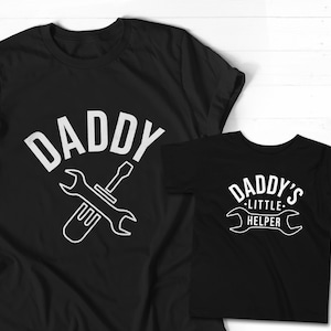 K&ouml;nnte beinhalten: Zwei schwarze T-Shirts mit wei&szlig;em Text. Das gr&ouml;&szlig;ere Shirt sagt "Daddy" mit einem Schraubenschl&uuml;ssel- und Schraubendreher-Design. Das kleinere Shirt sagt "Daddy's Little Helper" mit einem Schraubenschl&uuml;ssel-Design.