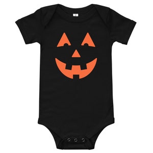 Puede incluir: Mono de beb&eacute; negro con un gr&aacute;fico de cara de calabaza naranja.