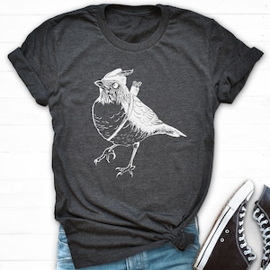 May include: Dark grey t-shirt with a white illustration of a bird wearing a hat and carrying a quiver of arrows. The bird is depicted walking. Black high-top trainers with white soles and laces are also visible.