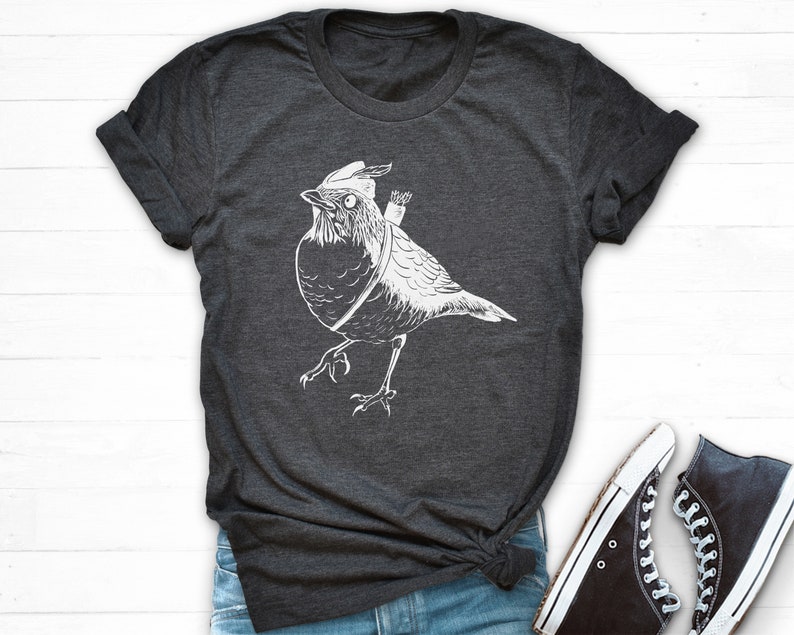 May include: Dark grey t-shirt with a white illustration of a bird wearing a hat and carrying a quiver of arrows. The bird is depicted walking. Black high-top trainers with white soles and laces are also visible.