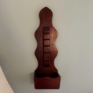 May include: A dark brown wooden wall-mounted rack featuring a vertical backboard with a wavy, scalloped edge. The backboard includes a central column of five small horizontal slats and a rectangular storage bin at the base with a wavy top edge.
