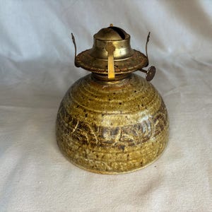 May include: A ceramic oil lamp base with a speckled olive-green glaze and a textured relief pattern around the lower section. The top features a brass burner assembly with two vertical prongs and a small circular adjustment knob on the side.