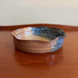 May include: A handcrafted ceramic bowl with a distinctive design. The bowl showcases a blend of brown, tan, and blue glazes. The rim has a gently undulating form, and the bowl features a double-banded pattern around the base. Ideal for serving or display.