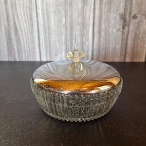 Pode incluir: Uma taça redonda de vidro transparente com um padrão de estrias verticais nas laterais. O recipiente possui uma tampa metálica dourada polida com superfície refletora e um pequeno puxador de vidro transparente no topo.