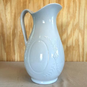 May include: A light blue ceramic pitcher with a glossy finish, featuring a curved handle, a wide pouring spout, and embossed oval panels decorated with delicate leaf patterns on the exterior surface of the rounded body.