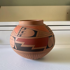 May include: A round, terracotta-colored pottery vessel with a wide opening. The vessel features geometric designs in black, red, and orange. The design includes zigzags, lines, and curved shapes. The pottery sits on a white surface.