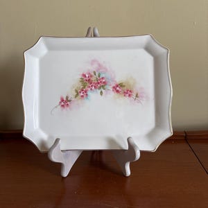 May include: A rectangular white serving tray with a scalloped edge and gold trim. The tray features a floral design with pink flowers and green leaves. The tray is displayed on a white stand.