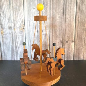 May include: A wooden carousel toy featuring three carved horses attached to a circular base. The structure includes a central pole topped with a yellow sphere, thin support strings, and small dark-colored finials above each horse.