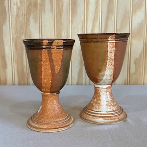 May include: Two ceramic goblets with a flared rim and a pedestal base. The left goblet features a mottled brown and green glaze with a textured finish. The right goblet displays a similar brown and green glaze with a prominent white vertical stripe.