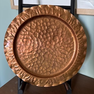 May include: A hammered copper tray with a textured surface and a ruffled edge. The tray has a circular shape and a warm, metallic color. The tray is likely used for decorative purposes.