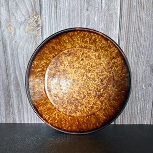 May include: A round ceramic plate featuring a mottled, variegated glaze in shades of deep amber, golden brown, and dark chocolate. The surface displays a textured, speckled pattern with a smooth, glossy finish and a dark brown rim.