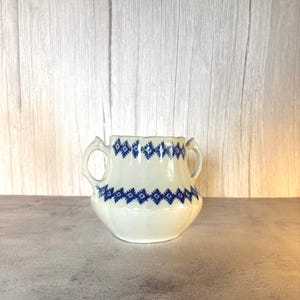 May include: A cream-coloured ceramic sugar bowl featuring two side handles and a scalloped rim. The exterior is decorated with two horizontal bands of a repeating blue geometric diamond pattern encircling the upper and middle sections.