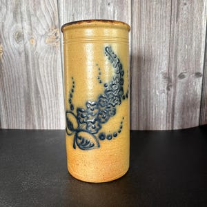 May include: A cylindrical stoneware crock with a tan, speckled finish and a dark brown rim. The exterior features a cobalt blue embossed floral design consisting of small flowers, leaves, and dotted patterns arranged in a vertical, flowing vine motif.