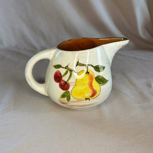 May include: A cream-colored ceramic creamer pitcher featuring a hand-painted design of a yellow pear and two red cherries with green leaves on the side. The pitcher has a rounded body, a curved handle, and a wide pouring spout with a brown rim.