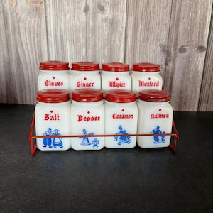 May include: A set of eight white milk glass spice jars with red metal lids, arranged in a red wire rack. Each jar features red text labels for Cloves, Ginger, Allspice, Mustard, Salt, Pepper, Cinnamon, and Nutmeg, with blue illustrations.