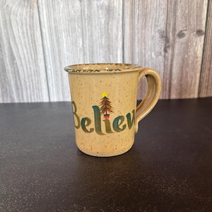May include: A beige speckled ceramic mug with a curved handle. The word Believe is printed in large green letters across the front, with a small, colorful illustrated Christmas tree featuring a yellow star replacing the letter i.