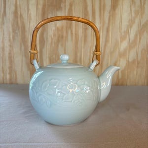 May include: A light blue ceramic teapot featuring a rounded body with an embossed floral pattern, a matching lid with a small knob, and a curved spout. The teapot includes a tall, arched handle made of natural brown bamboo with woven accents.