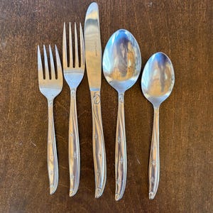 May include: A five-piece stainless steel flatware set including a small fork, a large fork, a dinner knife, a large spoon, and a small spoon. Each handle features a polished finish with engraved starburst accents and a curved, flowing line design.