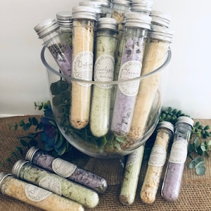 May include: A glass bowl filled with small glass tubes containing bath salts. The tubes have labels with different scents, including lavender, eucalyptus, and oatmeal mint. The bowl is sitting on a burlap surface with green succulents.