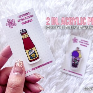 May include: Two 2-inch acrylic pins inspired by Filipino Jufran banana sauce. One pin features a cartoon bottle of banana sauce with the text "Banana Ketchup" and the other pin features a cartoon boba tea with purple tapioca pearls.