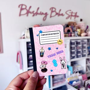 May include: A pink composition notebook with a glittery texture, decorated with stickers of a boba drink, donuts, a bunny, and a smiley face. The notebook has the words "Composition Notebook" and "Team Boba" printed on it.