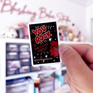 May include: A rectangular sticker with the words "YOU ROCK" in red, outlined in white, on a black background. The sticker also features a red strawberry graphic and the text "DON'T EVER CHANGE!" in white. The sticker is held by a hand.