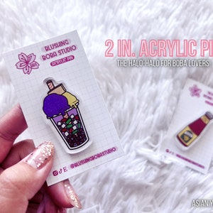 May include: Two 2-inch acrylic pins featuring a boba tea and a bottle of ketchup. The boba tea pin is purple, yellow, and green with a straw and boba pearls. The ketchup bottle pin is red with a yellow label. The pins are on white cards with the text "Blushing Boba Studio" and "2 in. Acrylic Pin The Halo-Halo for Boba Lovers!"