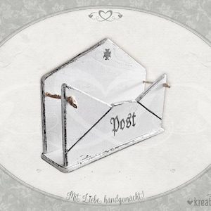 Könnte beinhalten: Weißer, hölzerner Briefkasten in Form eines Umschlags mit dem Wort "Post" auf der Vorderseite. Der Briefkasten hat eine abgenutzte Oberfläche und ist mit einer Schnur Schleife verziert.