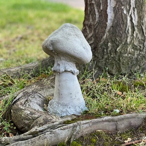 May include: A gray concrete mushroom garden statue with a textured surface. The statue is placed at the base of a tree.