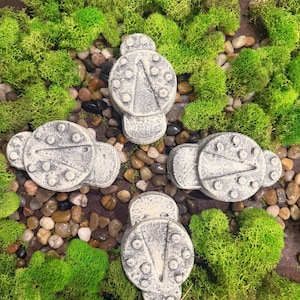 May include: Four gray, stone-like decorative objects with a circular top and a triangular design, surrounded by green moss and small pebbles. The objects have a weathered appearance, suggesting an aged or antique aesthetic.