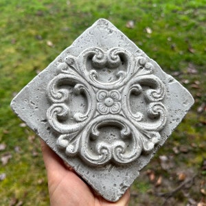 May include: A white concrete tile with a decorative, ornate, floral design. The tile has a rough, textured surface.