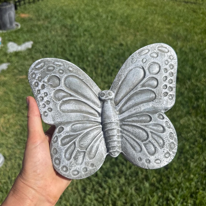 May include: A gray concrete butterfly sculpture with detailed wing patterns and a textured surface. The butterfly is held in a hand against a backdrop of green grass, showcasing its decorative design. This garden art piece is a unique outdoor accent.