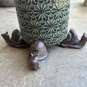May include: Three brown snail-shaped metal plant stand feet supporting a green and white ceramic planter with a geometric pattern.