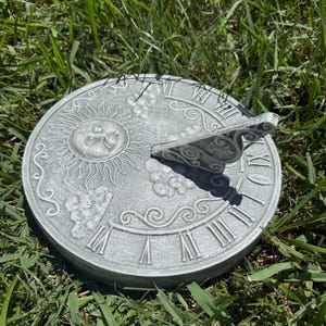 May include: A round, gray sundial with Roman numerals and a sun design. The sundial has a raised gnomon and is set in green grass. The sundial is a decorative garden accent.