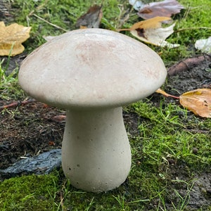 May include: A white concrete mushroom garden statue with a smooth, rounded top and a textured base. The statue is sitting on a bed of green moss.