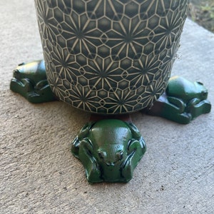May include: A dark green ceramic pot with a geometric pattern sits atop three dark green frog-shaped plant pot feet. The frog feet are arranged in a triangular pattern, providing support and a decorative touch to the plant pot.
