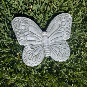 May include: A detailed, gray butterfly garden ornament with textured wings and a central body. The butterfly is resting on a bed of green grass, showcasing its intricate design and shape. The wings feature a pattern of circles and lines.