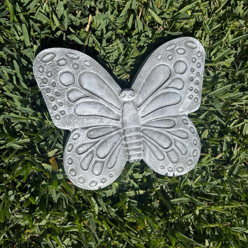 May include: A detailed, gray butterfly garden ornament with textured wings and a central body. The butterfly is resting on a bed of green grass, showcasing its intricate design and shape. The wings feature a pattern of circles and lines.