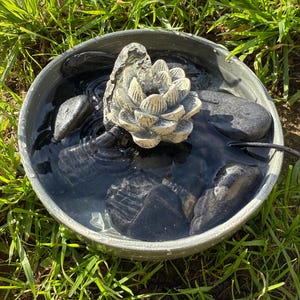 May include: A small, gray, circular water fountain with a white, ceramic lotus flower in the center. The fountain is filled with water and has black stones around the base.